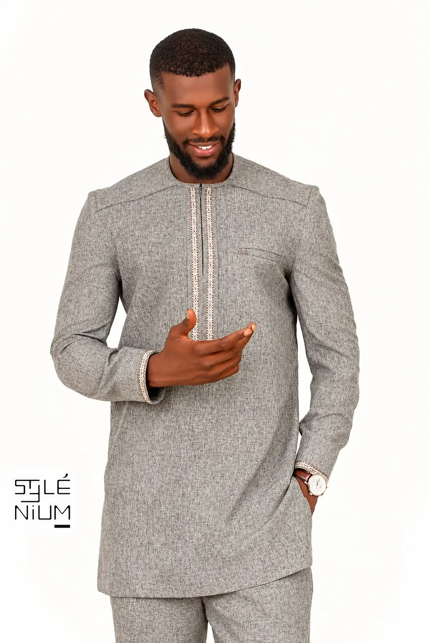 Homme souriant portant un ensemble tunique gris texturé avec galons brodés beige au plastron et aux poignets, création AMG STYLENIUM