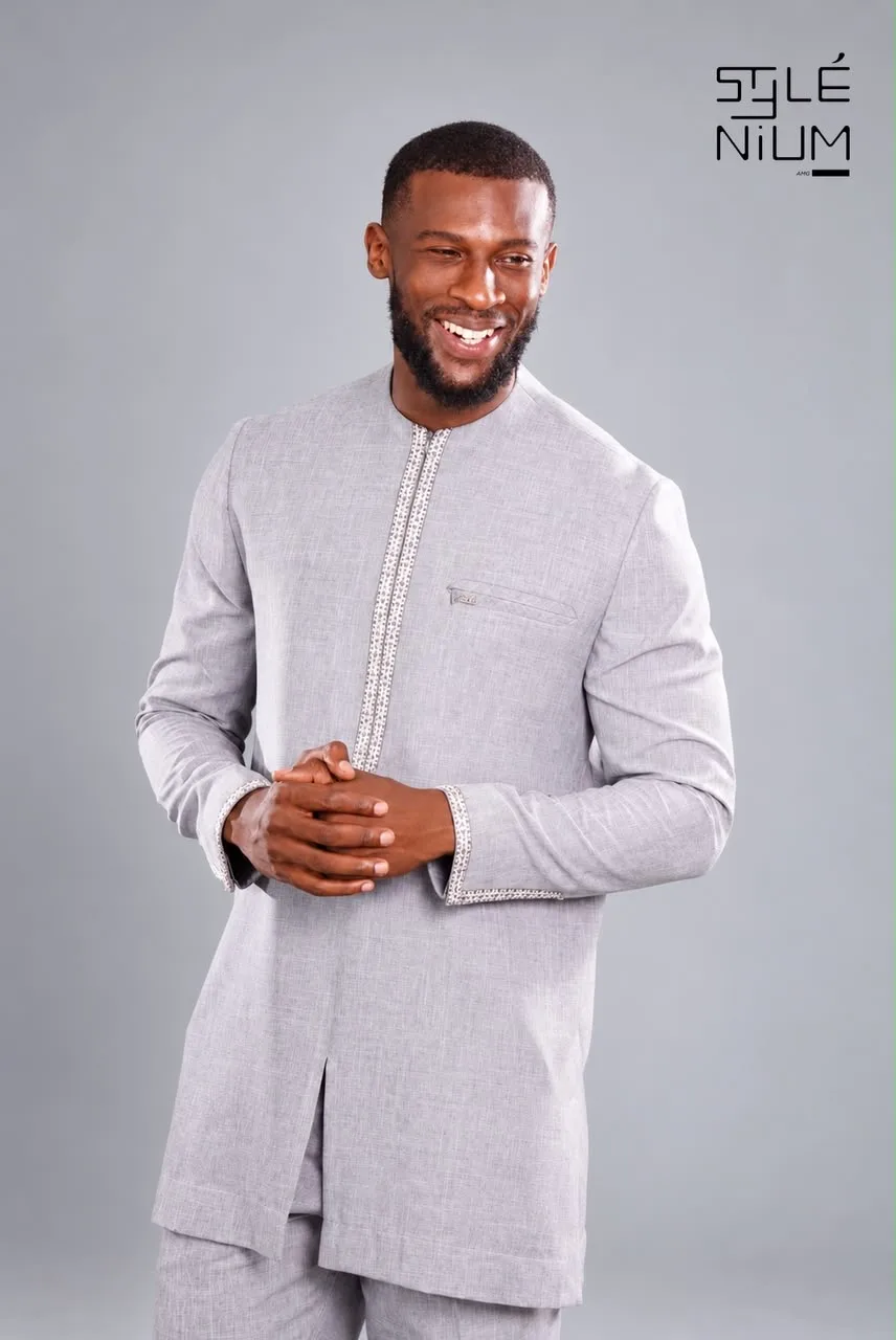Homme souriant portant une tunique gris chiné avec galons brodés à l'ouverture et aux poignets, création AMG STYLENIUM