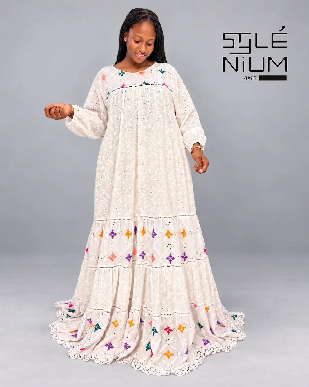 Femme portant une longue robe blanche ajourée à volants, ornée de petits motifs brodés multicolores et d'un ourlet festonné, création AMG STYLENIUM